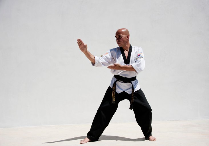 A person in a martial arts uniform and black belt performs a focused pose against a plain white background. They stand barefoot with one arm extended forward and the other bent across their body.