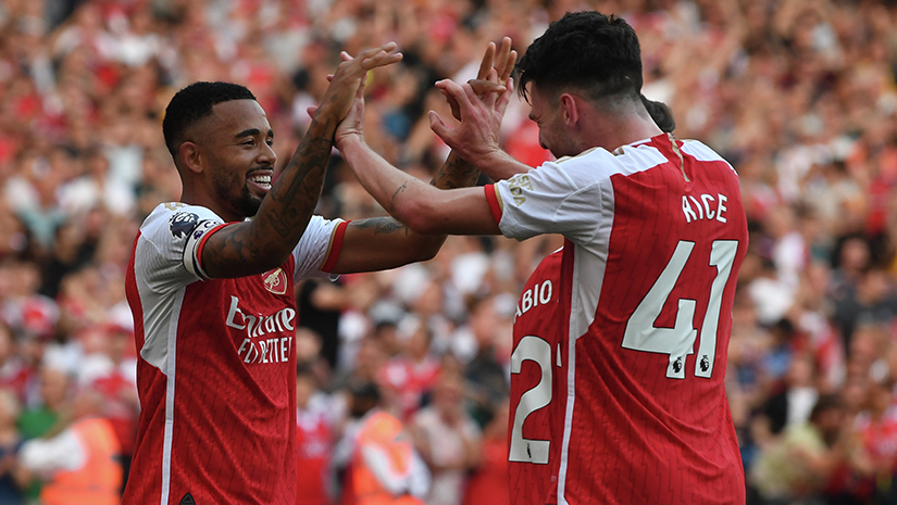 Three soccer players in red jerseys celebrate on the field, smiling and raising their hands. The crowd in the background is blurred, creating a lively atmosphere reminiscent of a thrilling Betway Arabia match.