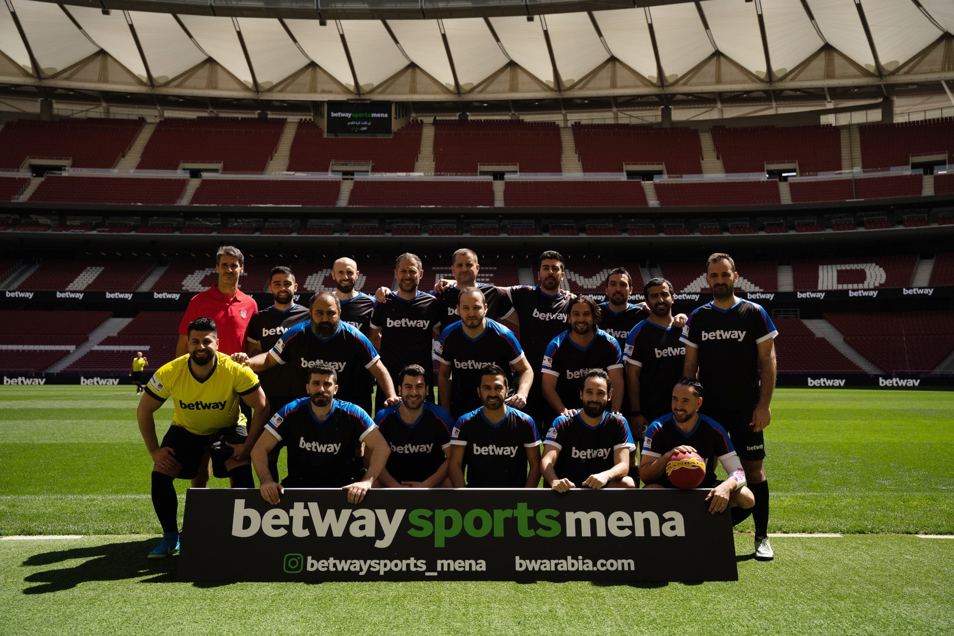 Un groupe dhommes en tenue de football posent sur un terrain de football dans un stade. Ils se tiennent derrière un panneau sur lequel est écrit « betwaysportsmena » et qui représente un ballon de football. Les sièges du stade sont visibles en arrière-plan.
