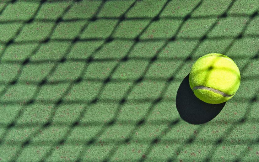 Une balle de tennis jaune vif projette une ombre sur la surface verte d'un court. Le premier plan est traversé par l'ombre d'un filet de tennis, formant un motif en treillis rappelant la disposition structurée que l'on retrouve dans les arènes sportives Betway à travers l'Arabie.