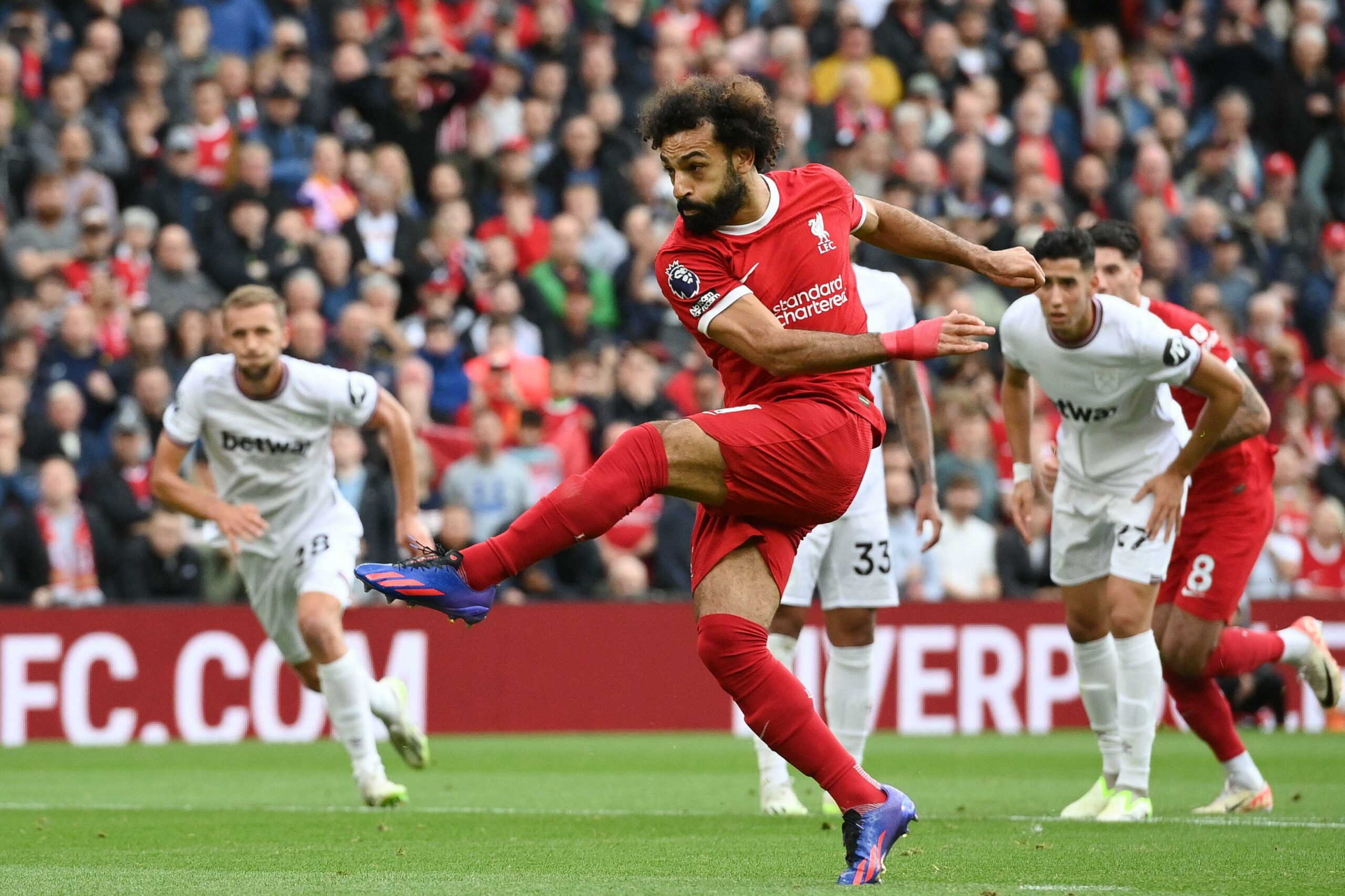 A soccer player in a red jersey expertly kicks the ball during a match, with players in white jerseys and spectators eagerly watching from the background, adding to the thrilling atmosphere of this Betway Arabia-sponsored event.