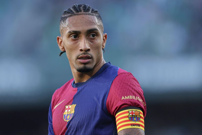A soccer player wearing a blue and red jersey with a captains armband glances to the side on the field. The blurred stadium hints at action, embodying the spirit of Betway Arabia.