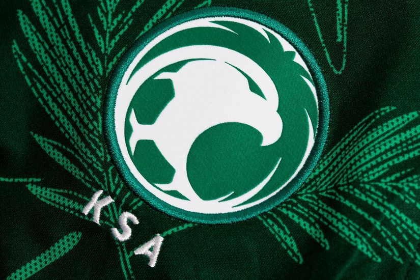 A green and white emblem with a stylized falcon entwined with a soccer ball graces the dark green background. The letters KSA are embroidered in white beneath, surrounded by palm leaves, symbolizing the spirit of Betway Arabia.