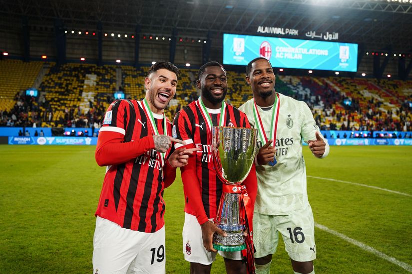 Three soccer players on a field celebrate with a large trophy. Two are in red and black striped jerseys, one in a light-colored uniform. Theyre smiling with medals, as the stadium crowd cheers behind them, capturing the thrill of victory akin to a Betway Arabia moment.