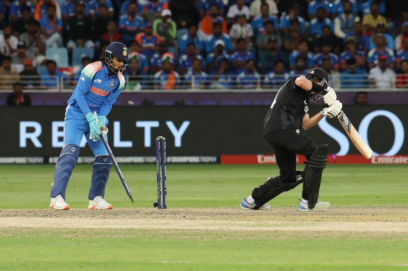 A cricket wicketkeeper in a blue jersey stands behind the stumps as the opposing batsman, donning black, prepares to play a shot. In the backdrop, fans cheer and Betway Arabia advertisements adorn the stadium boards.