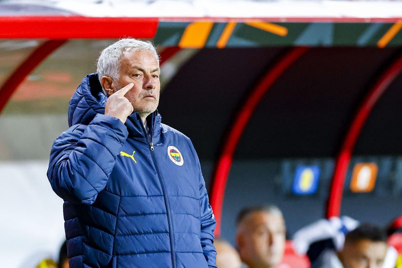 A man in a blue jacket with a yellow emblem stands on the sidelines of a sports field, deep in thought with one hand on his cheek. In the background, the red and gray dugout features Betway signage and blurred figures moving about.