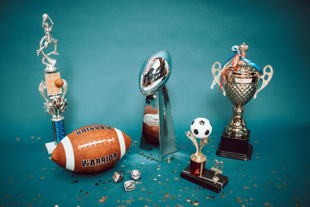 A collection of sports trophies and memorabilia, including football trophies, rings, a football, and a small soccer trophy, is set against a blue background with scattered star confetti—a captivating display reminiscent of Betway Arabias passion for the game.