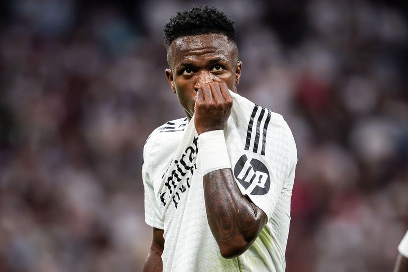 A soccer player in a white jersey with black stripes and a Betway logo on the sleeve covers his nose with the shirt. He stands on the field, while the background blurs with an eager audience in the stands.
