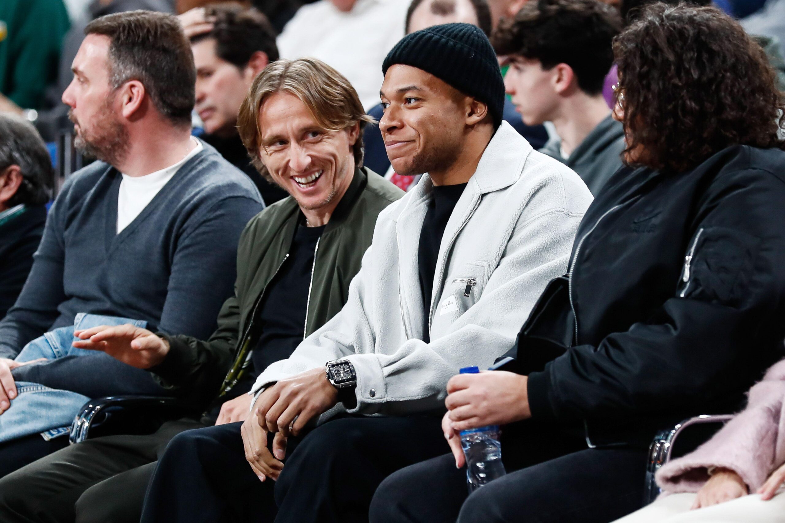 Four people are seated in a row at what appears to be a sports event. They are engaged in conversation and smiling. The man on the left gestures with his hand. The audience can be seen in the background, slightly out of focus.