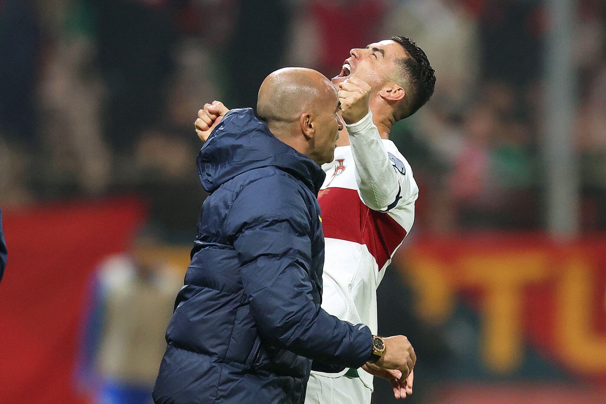 يحتفل لاعب كرة قدم يرتدي قميصًا أبيض وأحمر بذراعيه مرفوعتين وفمه مفتوحًا، بينما يقف شخص آخر يرتدي سترة زرقاء بالقرب منه. تلتقط الخلفية النابضة بالحياة البهجة في تلك اللحظة، تمامًا مثل الإثارة التي توفرها عروض Betway Arabia لعشاق الرياضة.