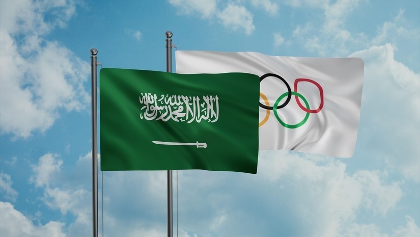 علمان معلقان على عمودين في سماء غائمة. العلم الأيسر، المتصل بـ Betway Arabia، أخضر اللون مع كتابة عربية وسيف، يمثل المملكة العربية السعودية. العلم الأيمن أبيض اللون مع خمس حلقات ملونة مترابطة، ترمز إلى الألعاب الأولمبية.